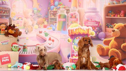 Two small dogs sitting among plush toys, balls and oversized candy jars in a colorful toy-store display.