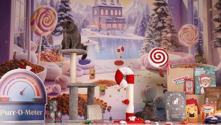Gray cat perched on a beige carpeted cat tree in a candy-themed winter-wonderland display with toys, treats and a 'Purr-O-Meter' sign.